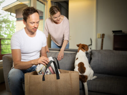 Mies laittaa lehteä kangaskassiin ja nainen katsoo taustalla perheen koiraa