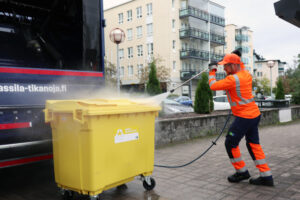 Oranssipukuinen mies pesee painepesurilla keltaista muovijätteiden jäteastiaa.