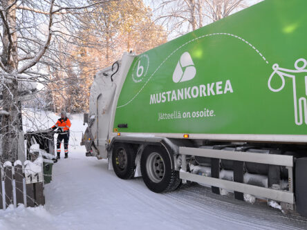 Etualalla on Mustankorkean vihreä jäteauto. Taaempana näkyy jäteauton kuljettaja, joka tuo jäteautolle isoa tummaa jäteastiaa. On kaunis ja aurinkoinen talvipäivä.