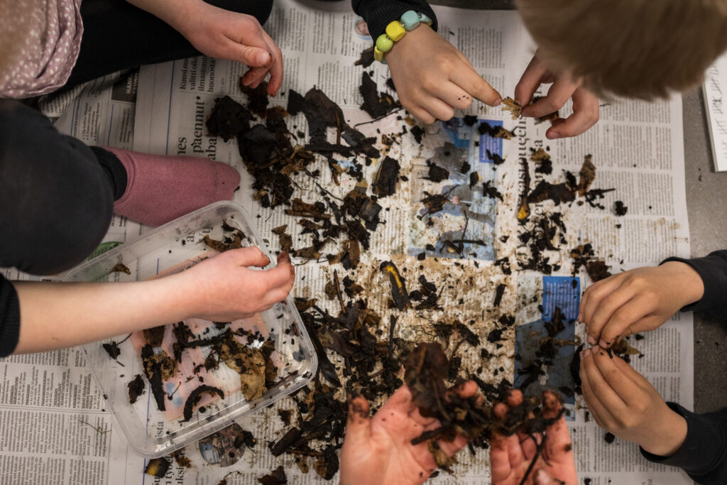 Sanomalehtien päällä on multaa ja matoja. Kuvassa näkyy lasten käsiä.
