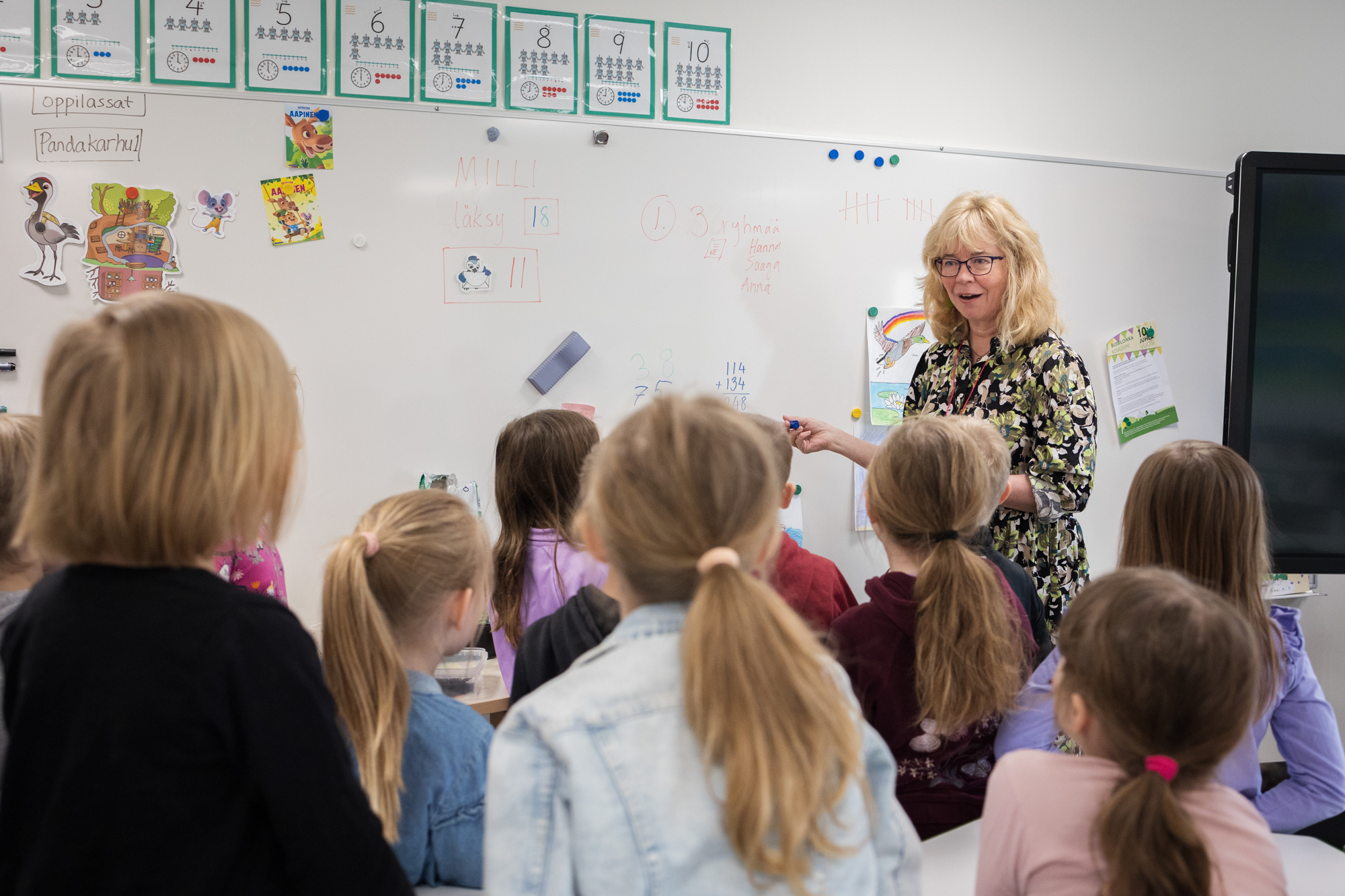 Vaaleahiuksinen opettaja kertoo luokalle jotain. Lapset ovat kameraan selin.