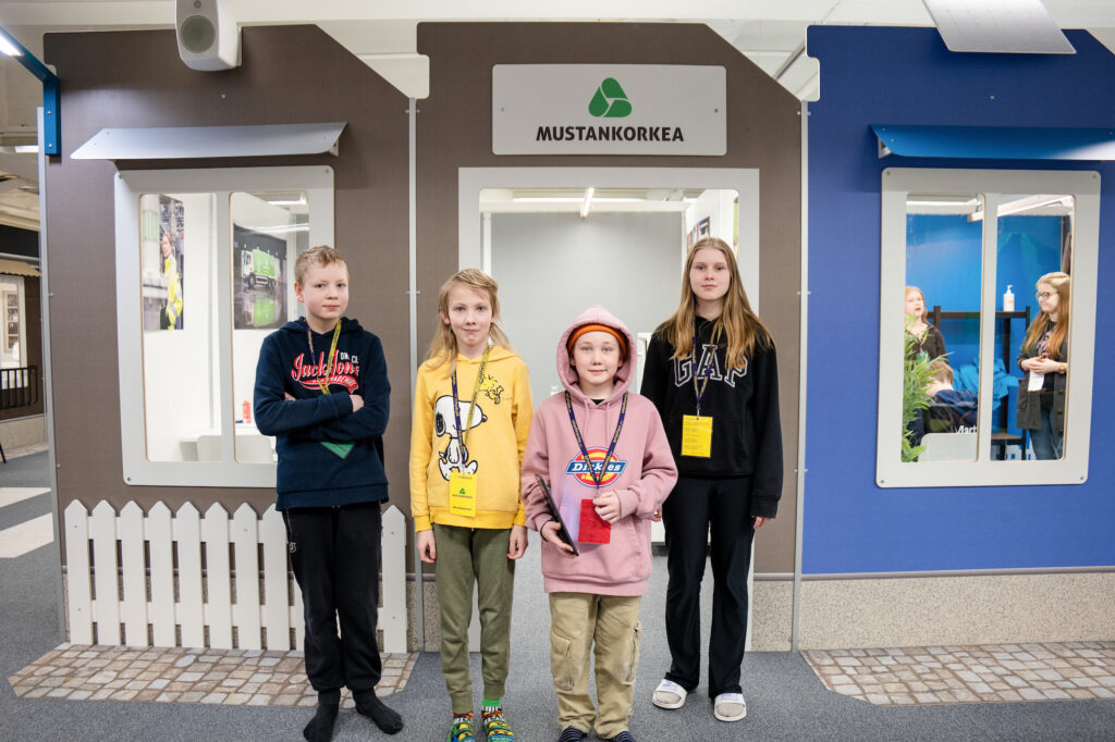 Neljä oppilasta seisoo rivissä Mustankorkean Yrityskylä-pienoisyrityksen edessä.