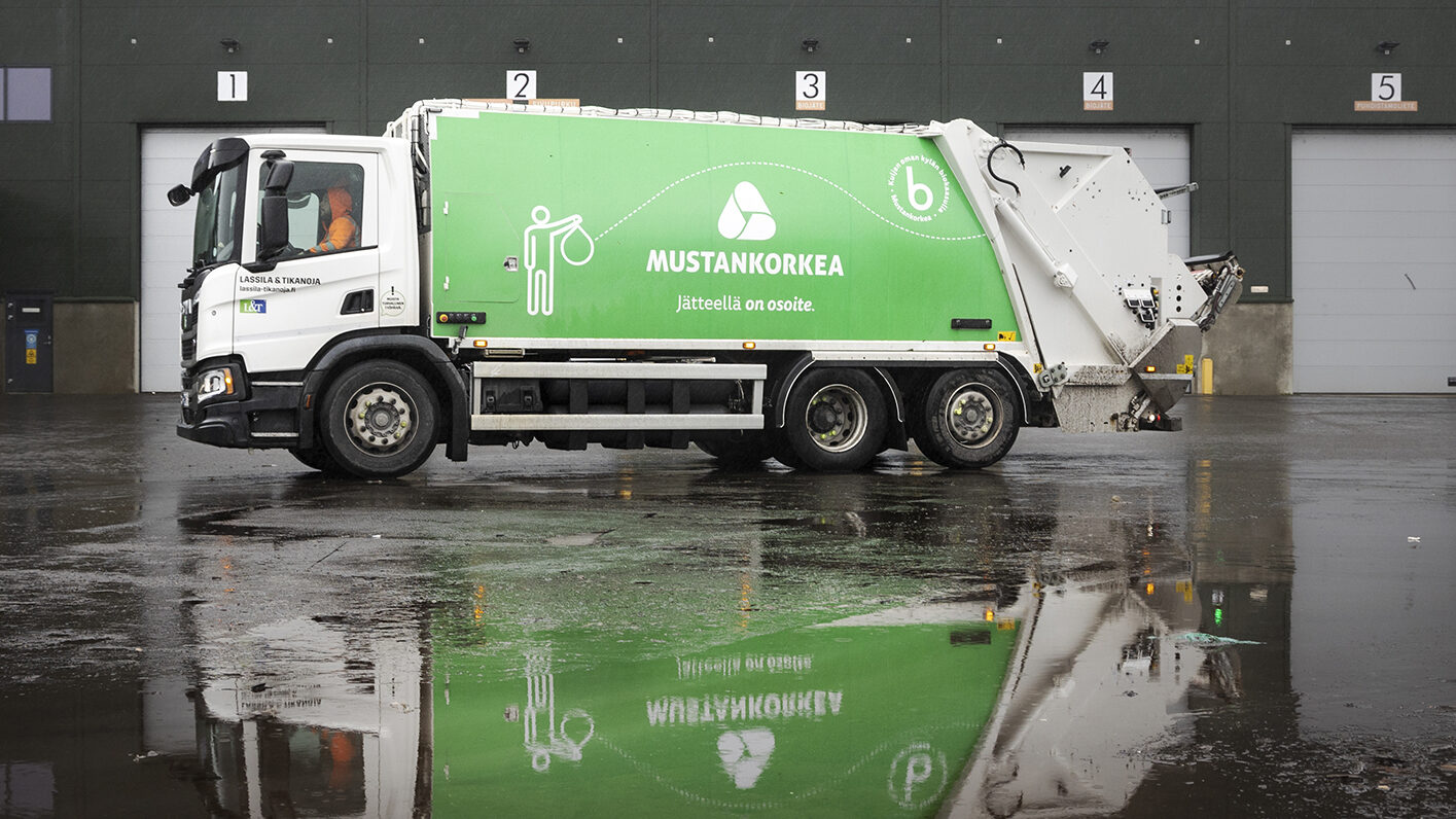 Vihreä Mustankorkean jäteauto on parkissa biokaasulaitoksen ovien edessä. On sateinen päivä ja vesilammikosta näkyy heijastus autosta.