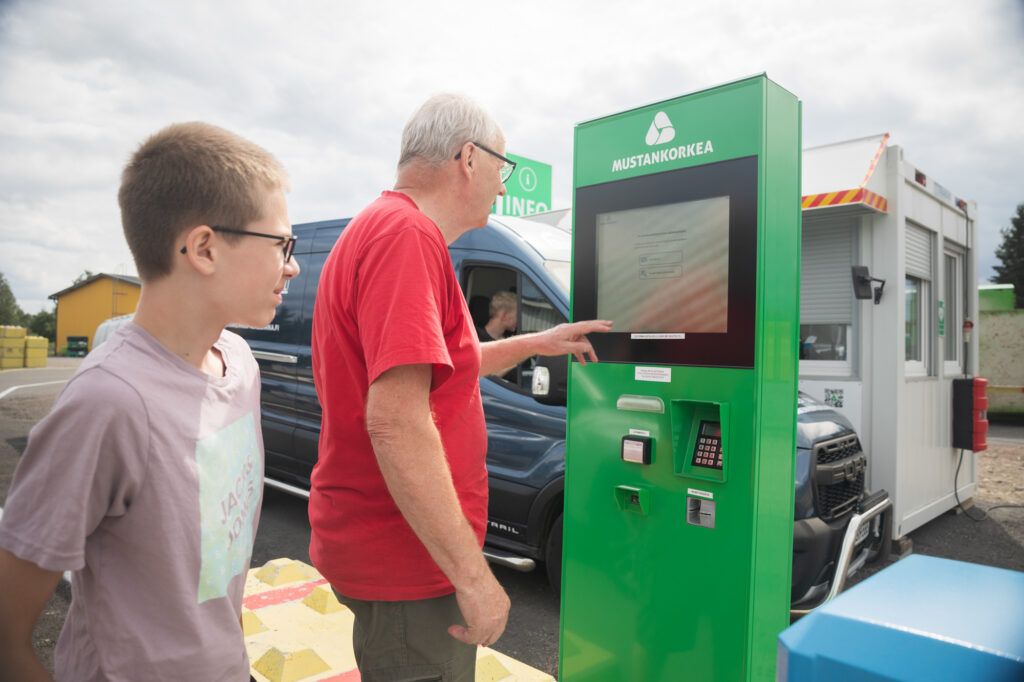 Punapaitainen vanhempi mies asioi vihreällä automaatilla. Hänen vieressään seisoo nuorukainen. Automaatin takana on pakettiauto peräkärryn kanssa infopisteellä.