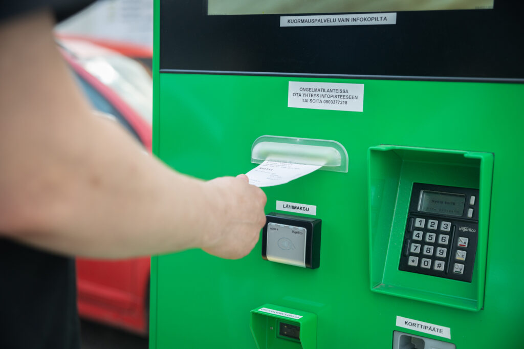 Vihreästä automaatista tulee kuitti, jota käsi vastaanottaa. Oikealla on korttimaksun numeronäppäimet.