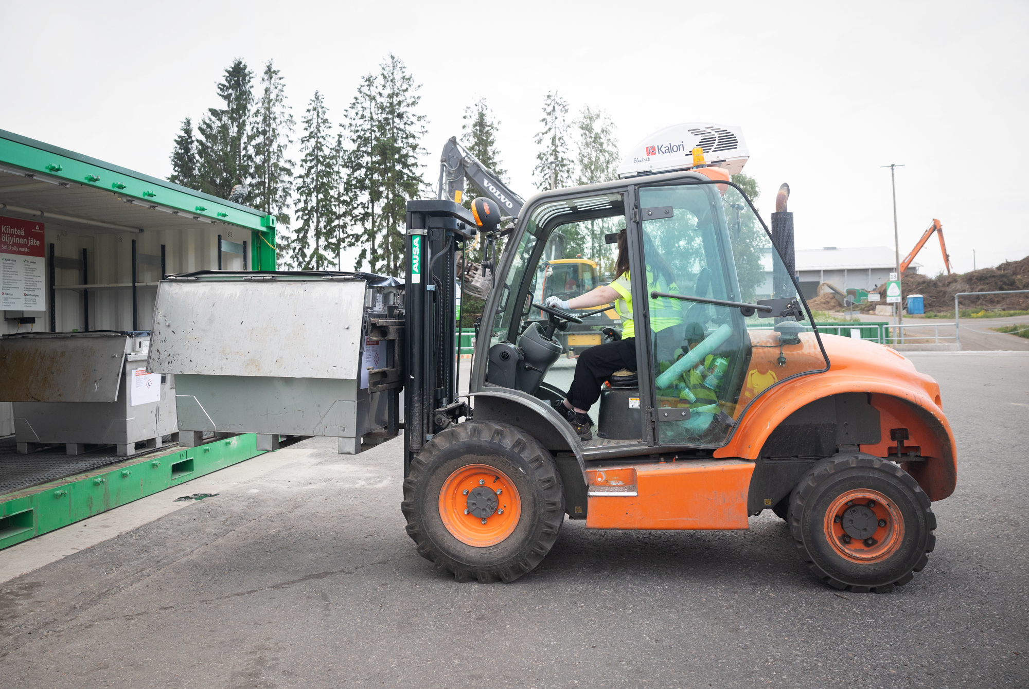 Oranssilla trukilla on kyydissä harmaa iso metallinen laatikko. Kuljettajana on pitkähiuksinen nainen, jonka kasvoja ei näy.