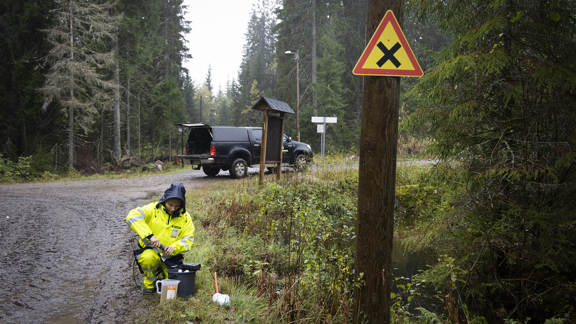 Huomiovaatteet päällä oleva nainen on ottamassa vesinäytteitä. Hänellä on edessään erilaisia pulloja ja sanko. On sateinen päivä.