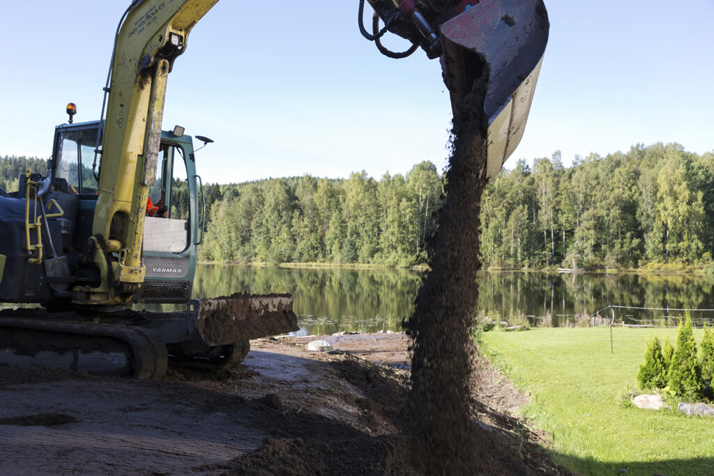 Kaivinkone levittää multaa ja taustalla näkyy kaunis järvimaisema.