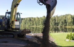 Kaivinkone levittää multaa ja taustalla näkyy kaunis järvimaisema.