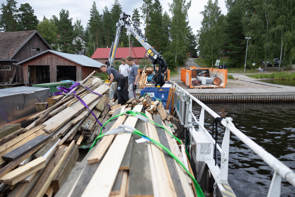 Laivan kannella on liinoilla käytettynä puujätettä. Takana näkyy oransseja jätelavoja.