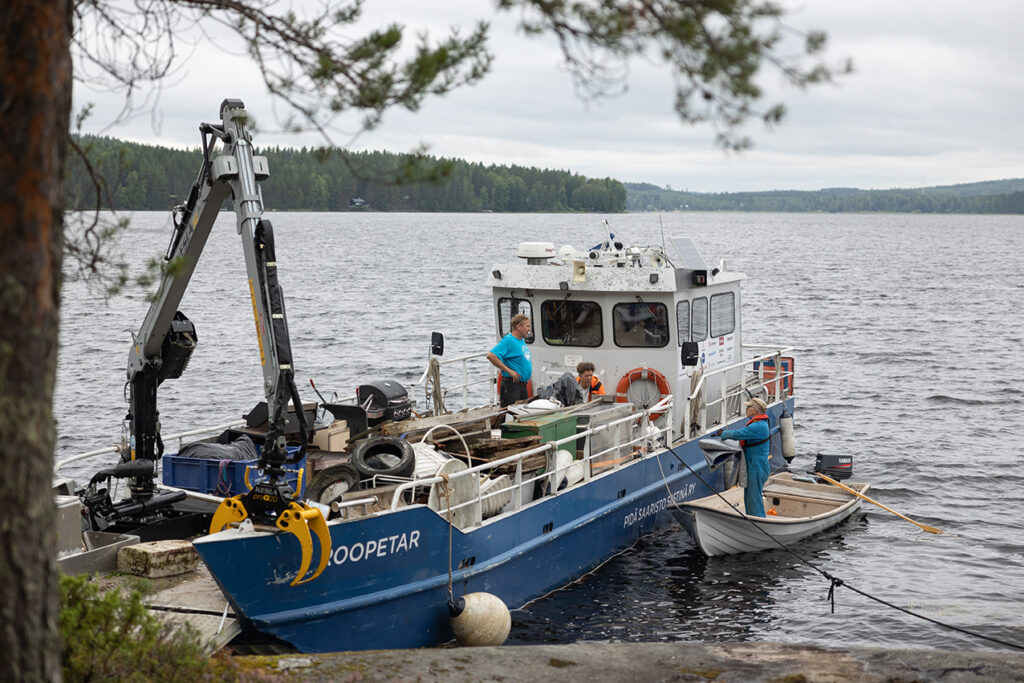 Kuvassa näkyy sinivalkoinen huoltolaiva ja vieressä soutuvene, joka ojentaa laivaan jotain. Sää on hieman pilvinen.