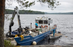 Kuvassa näkyy sinivalkoinen huoltolaiva ja vieressä soutuvene, joka ojentaa laivaan jotain. Sää on hieman pilvinen.