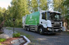 Mustankorkean jäteauto on jossain pihalla. Edessä näkyy istutuksia ja ulkovalaisin, takana jokin pyörähirsirakennus ja hieman lasten pinnasänkyä.