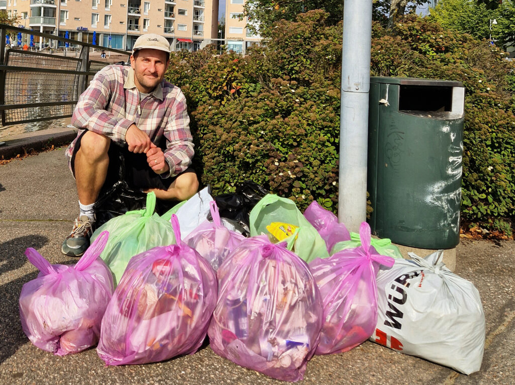 Mies katsoo kameraan ja hymyilee. Edessä on lähemmäs kymmenen pussia roskia pääsääntöisesti pinkeissä ja vihreissä roskapusseissa.