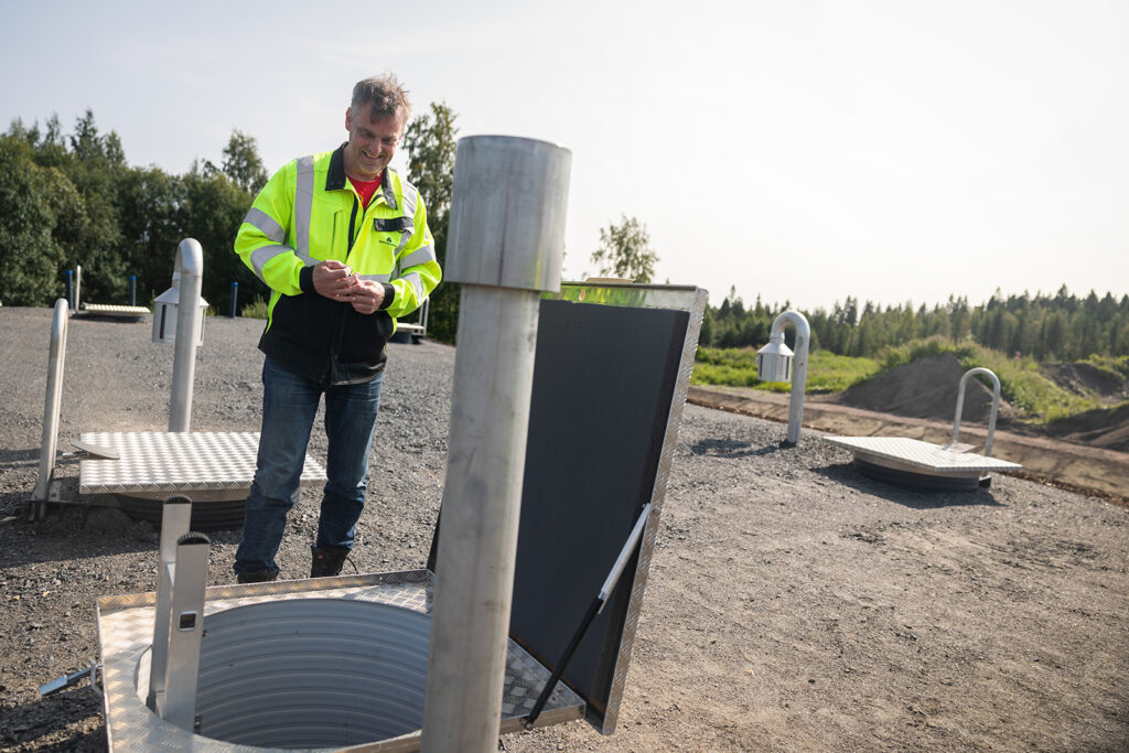 Keltainen huomiotakki päällä oleva mies katsoo alaspäin jonkinlaisesta maan alle menevästä teräsputkesta alas ja hymyilee. Takana näkyy alempana metsää.