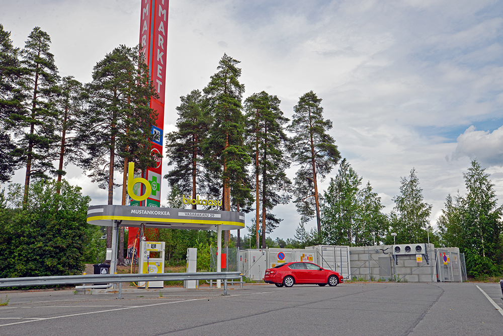 Tankkausaseman ylhäällä on b-kirjain ja takana näkyy punainen Citymarketin pylväs. Tankkaamasta on lähtenyt punainen auto.