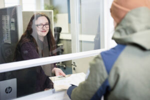 Silmälasipäinen ja tummahiuksinen nainen ojentaa jotain lappua asiakkaalle, josta näkyy vain selkä. Nainen hymyilee kauniisti.