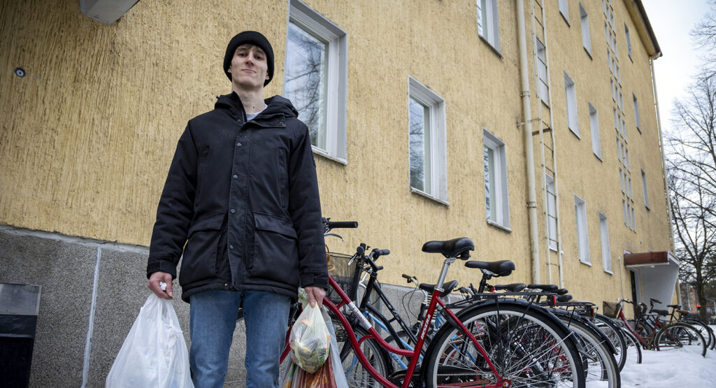 Nuori mies kantaa roskapusseja. On talvi ja ulkona on lunta. Takana näkyvä kerrostalo on keltainen ja sää on melko harmaa. Mies hymyilee pienesti.