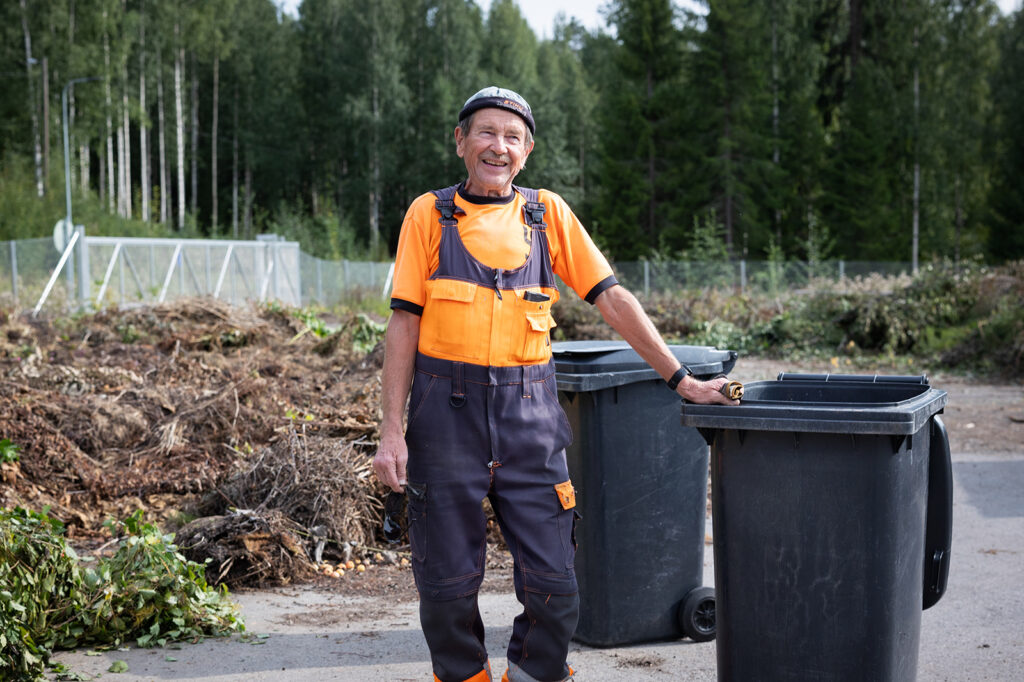 Mies nojaa jäteastiaan ja hymyilee. Takana näkyy puutarhajätettä.