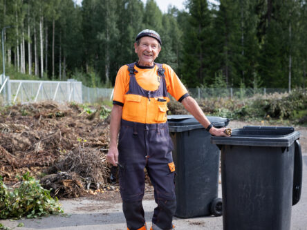 Mies nojaa jäteastiaan ja hymyilee. Takana näkyy puutarhajätettä.