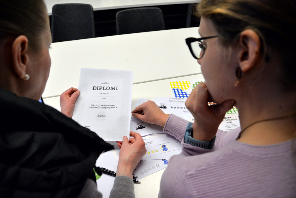 Kaksi naista tutkii toisen kädessä olevaa harmaata diplomia. Naiset on kuvattu heidän selän takaa.