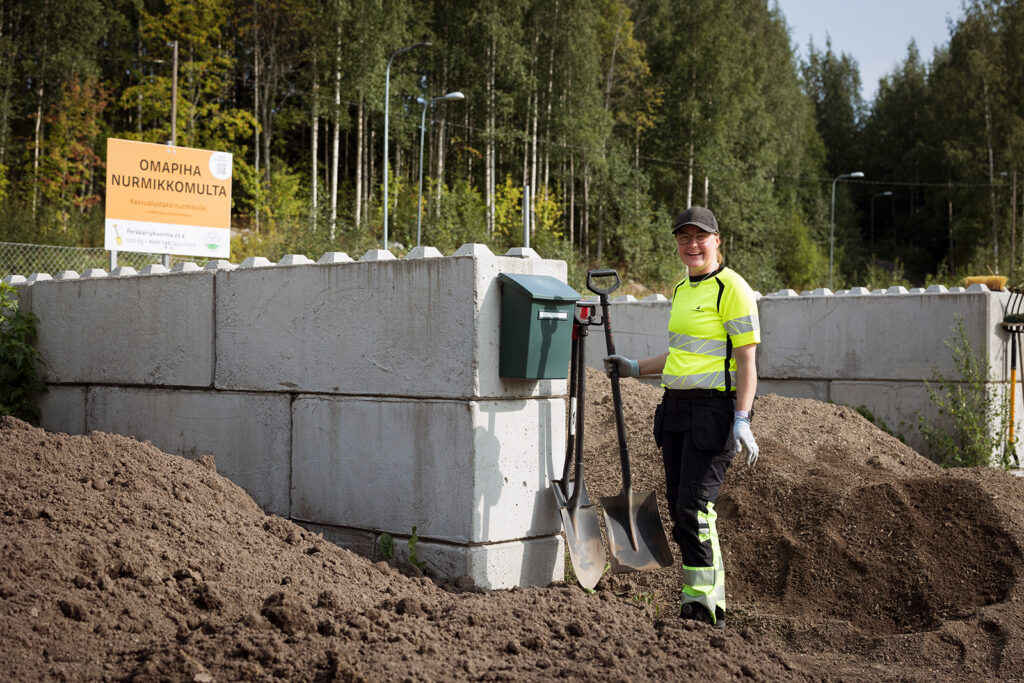 Huomiovaatteissa oleva nainen pitää lapiota ja katsoo nauraen kameraan. Sivuilla näkyy kaksi multaloossia.