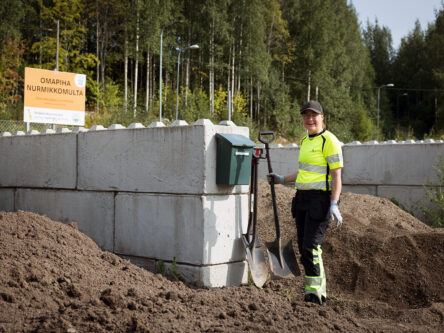 Huomiovaatteissa oleva nainen pitää lapiota ja katsoo nauraen kameraan. Sivuilla näkyy kaksi multaloossia.