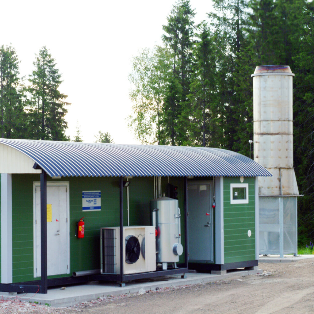 Jokin vihreä junanvaunun näköinen rakennus, jonka vieressä on metallinen korkea piippu tai vastaava. Kuva on hieman vanha.