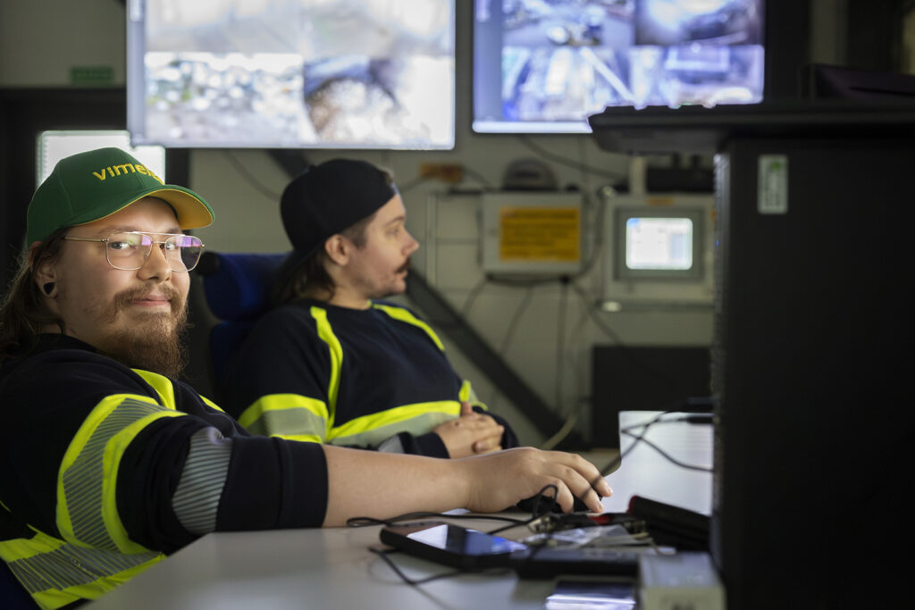 Kaksi miestä istuu monitoreiden edessä. Toinen katsoo hymyillen kameraan, toinen katsoo toisaalle. Takana näkyy lisää näyttöjä.