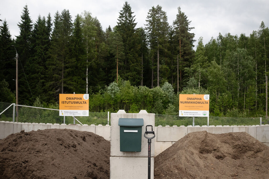 Kaksi multakasaa on erotettu toisistaan betonielementeillä. Niiden keskellä on vihreä postilaatikko. Multakasojen takana on kyltit, joissa toisessa lukee Omapiha Istutusmulta ja toisessa Omapiha Nurmikkomulta.